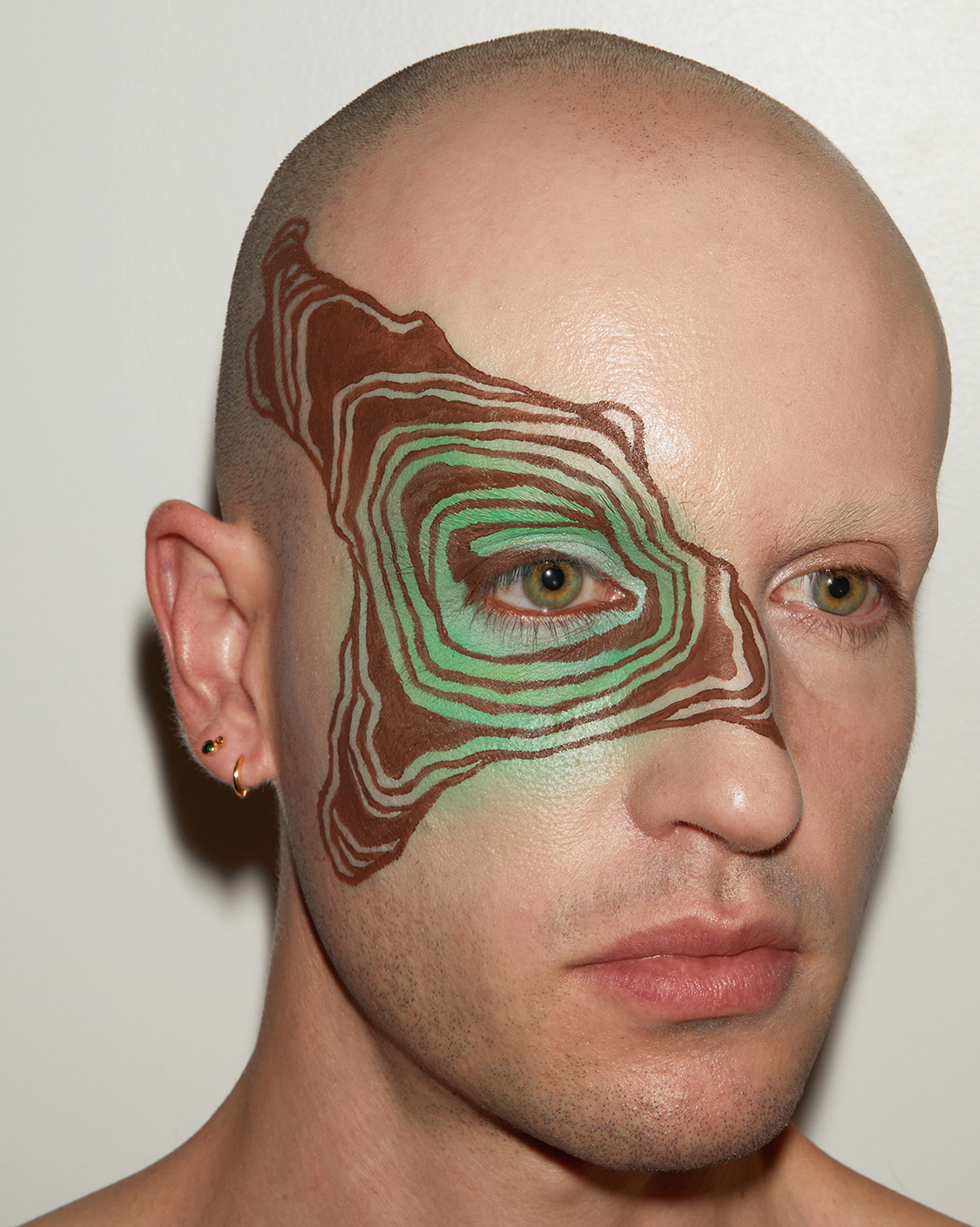 Close up portriat of Jesse at 3/4 angle with graphic brown liner make up in concentric shapes extending out from one eye against a grey background